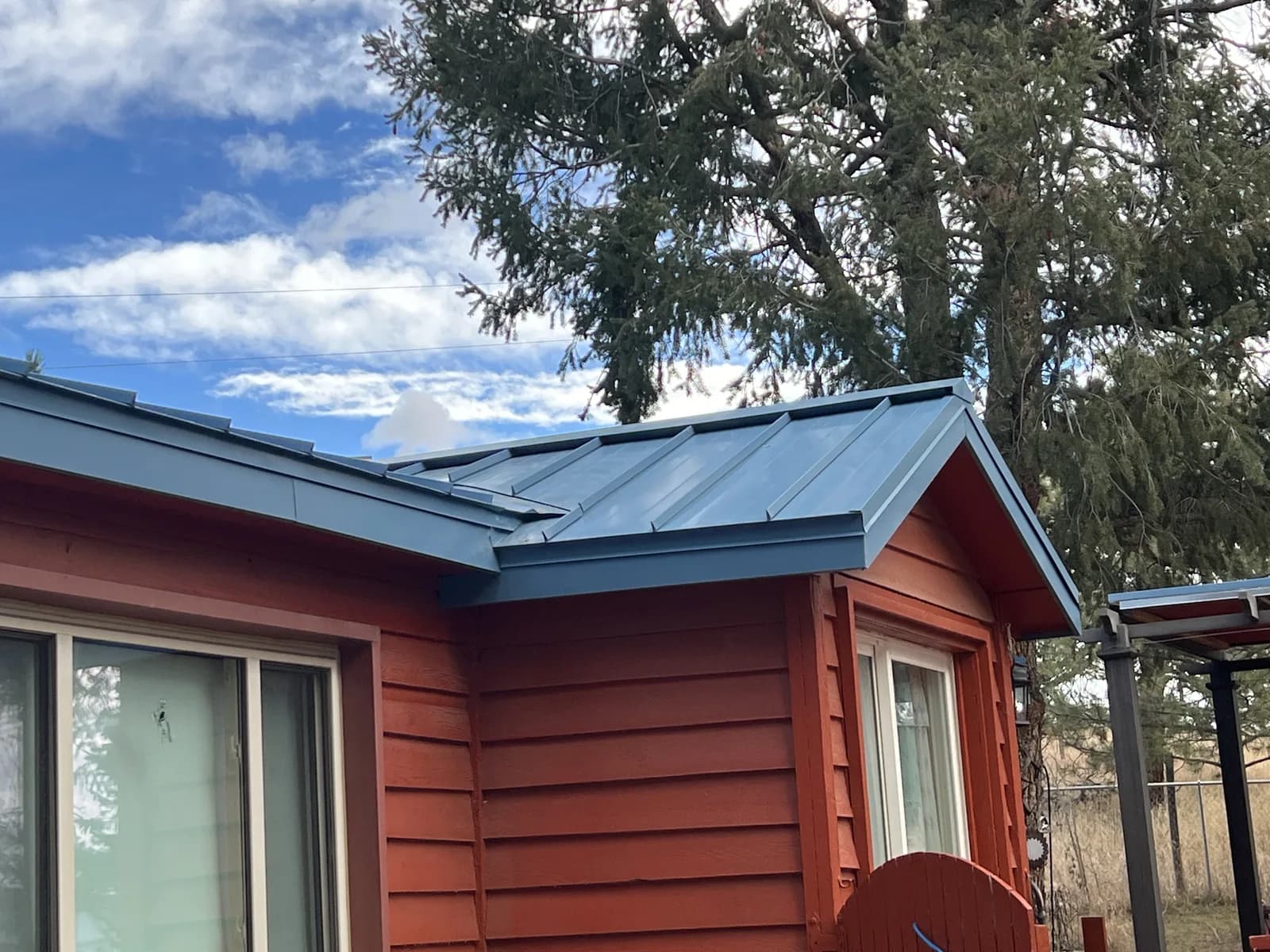 Blue-gray standing-seam roof panels above red siding and windows beneath a tree
