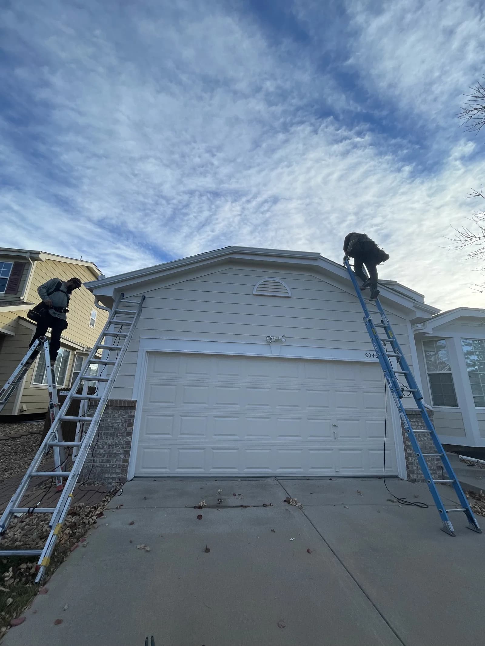 Two workers on access ladders beside white siding and the roofline above a garage